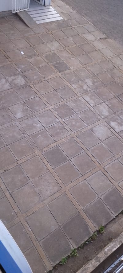 The scene depicts an outdoor paved area in Ngong ward, Kenya, viewed from an elevated perspective. The primary surface consists of numerous square, light grey concrete pavers arranged in a grid, separated by narrower, lighter-colored concrete strips that create a distinct pattern. Minor wear and discoloration are visible on some pavers.
In the upper-left quadrant, a white metal gate or railing, partially open, is visible, leading to an entrance. Below this, a concrete accessibility ramp descends, bordered by a section of light-colored horizontal strips, possibly steps or a striped pathway. The wall behind this entrance is light-colored.
Towards the upper-right, the paved area transitions into a section of darker, smaller, rectangular bricks laid in a herringbone pattern. The bottom-right edge of the main paved area borders a strip of bare earth with sparse green vegetation, specifically small plants resembling weeds. A concrete curb or edge is present along this boundary. The lower-left foreground shows a partial view of a blue and white object, possibly a vehicle part or another railing. A small, white, rectangular piece of debris is located on one of the central pavers. No individuals, discernible actions, or interactions are present in the frame. The lighting is even, suggesting an overcast condition.