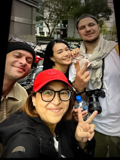 The image captures a group of four individuals posing for a selfie on a street in Taipei, Taiwan. In the foreground, a person wearing a red baseball cap and glasses smiles at the camera, holding a bottle of water. To their left, a person with short brown hair and sunglasses peeks into the frame. Behind them, a woman with long dark hair smiles and makes a peace sign. To the right, a man with a beard and a scarf draped around his neck also makes a peace sign, with a camera visible on his shoulder. They are standing in front of a row of motorcycles parked along a sidewalk, with a building and trees visible in the background, suggesting an urban setting. The overall atmosphere appears casual and friendly.