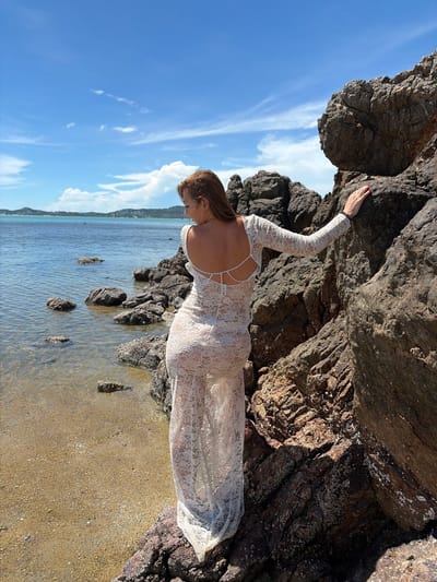 A woman in a white lace dress stands on rocky terrain near the water's edge, with her back to the camera. She is touching a large rock to her right. The water is calm and clear, with small waves lapping at the shore. In the distance, a coastline with buildings and vegetation is visible under a blue sky with scattered clouds. The scene appears to be a tropical beach.