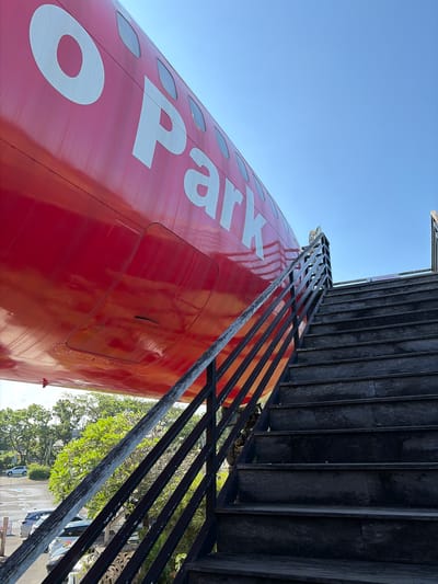 The image shows a red airplane fuselage, with the partial word "Park" visible in white lettering. A black wooden staircase with a metal railing leads upwards towards an opening in the plane. The stairs are worn and appear to be made of dark wood. Above the stairs, a sign with text in a foreign language is affixed to the railing. The sky is clear and blue. In the background, there are green trees and parked cars, suggesting an outdoor location. The context provided indicates this scene is in Blahbatuh, Indonesia.
