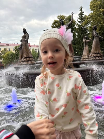 In this close-up, low-angle photograph, a young child, possibly a toddler, is captured from the chest up, positioned in the foreground. The child wears a light-colored, long-sleeved shirt adorned with a pattern of pink flowers and green stems. A knitted white cap with a pink flower embellishment sits on their head. The child's face is turned towards the camera, with an open mouth, suggesting they are speaking or laughing. Their eyes are visible, and their blonde hair is slightly tousled.
Behind the child, a fountain is actively spraying water, creating a dynamic and misty effect. Some of the water appears to be illuminated with purple and blue lights. In the background, several bronze statues of women in traditional attire are visible, some partially obscured by trees and water. These statues are likely part of a monument. The backdrop also includes a building with a red roof and a cloudy sky, hinting at an outdoor setting. The overall scene evokes a playful and joyful moment, possibly during a day out at a park or public square in Ufa, Russia. The time of day appears to be daytime, and the weather is overcast. There is no visible text in the image.