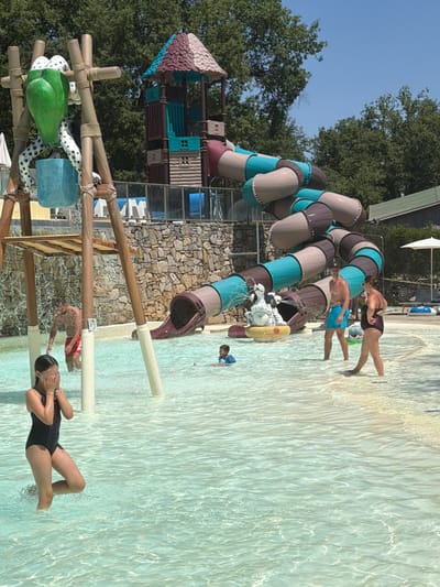 The image depicts a sunny day at a water park, identified as Acquapendente di Cavriglia in Italy. In the foreground, a young girl in a black swimsuit stands in shallow water, covering her eyes with her hands. Behind her, a man in red swim trunks is partially visible, also in the water, with water spraying from a structure above.
Further back, a water slide complex with multi-colored tubular slides dominates the scene. A man in blue swim trunks and a woman in a dark swimsuit stand near the exit of one of the slides. A small child in a blue bathing suit is also in the water near the slides. A decorative sculpture of a dog is positioned at the entrance to one of the slides. The water park features a stone wall in the background and lush green trees surrounding the area. The water is clear and shallow, with sunlight reflecting off its surface.