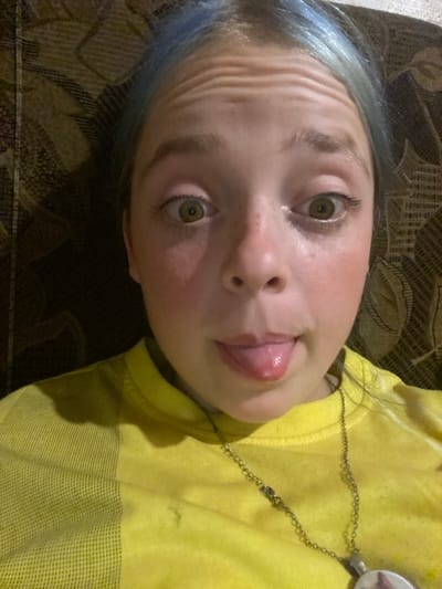 This is a close-up, Dutch angle, low-angle shot of a young person lying down. The person has light skin, green eyes, and blue-tinted hair. They are sticking their tongue out and have a playful expression. Their forehead is creased. They are wearing a bright yellow shirt with a striped pattern on the lower portion and a thin metal chain necklace with a round pendant. The background appears to be a brown couch with a textured pattern. The lighting suggests it's indoors, possibly in the evening. No visible text is present in the image. The location context is Nova Praha, Ukraine.