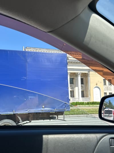 The image, taken from inside a vehicle looking out, captures a street scene in Chelyabinsk, Russia. A large truck with a blue tarp covering its cargo dominates the left side of the frame. Behind the truck, a yellow building with classical architectural elements, including columns and arched windows, is visible. The sky is clear and blue. A side-view mirror reflects a white van with a police light bar on its roof, suggesting a law enforcement presence. The foreground shows the interior of the vehicle, including the dashboard and door panel. The street is paved, and there is some greenery in front of the building. The overall impression is a snapshot of urban transit on a sunny day.