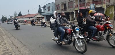The image depicts a busy urban street scene in Goma, Democratic Republic of the Congo, under an overcast sky. The foreground features a paved, grey asphalt road with a concrete curb and sidewalk visible on the left.

Two motorcycles dominate the central and right foreground, both moving towards the viewer. The motorcycle on the left is a red standard model with silver and chrome components, including a prominent blue-tinted headlight. It carries two male occupants: the front rider wears a grey hooded jacket and blue jeans, while the rear passenger wears a white hooded sweatshirt and a dark vest.

Immediately to the right, a second red motorcycle, modified with a wider rear track, appears to be a three-wheeled mototaxi. The male rider wears a dark jacket, blue jeans, and a yellow helmet. His female passenger, seated behind him, is dressed in a dark garment and holds a light-colored bag.

In the mid-ground and background, additional motorcycles and a white sedan are visible on the road. The right side of the street is lined with multi-story concrete buildings featuring balconies with metal railings and various commercial signs, including a prominent blue one and a red and white striped umbrella. The left side of the street features lower, single-story structures with reddish-brown roofs and sparse trees. Several other individuals are present in the background, interacting near the commercial establishments.