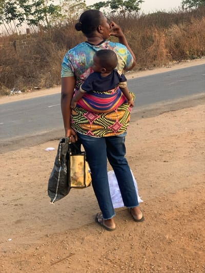 A woman stands on a road's edge, holding a child secured on her back with a brightly patterned cloth. The child wears a dark shirt. The woman has on a tie-dye shirt and jeans. She carries a bag over her shoulder. The background shows dry vegetation and a road. The scene captures an everyday moment, possibly a walk along the road.