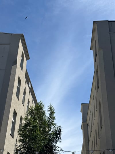 The media file captures an upward-angle shot of modern architecture against a clear blue sky with wispy white clouds. Two buildings are prominently featured, one above the other and angled towards each other, creating a V-shape in the frame. The upper building's underside is visible, revealing a structured facade with rectangular indentations. The lower building shows multiple windows, some rectangular and some with rounded tops, along with a satellite dish. A lone bird is silhouetted against the sky in the lower left quadrant. A tree with dense green foliage overhangs the lower building from the right. String lights with small white spherical bulbs are visible hanging down from the upper structure on the right side. The lighting suggests it is daytime, likely late morning or early afternoon, with the sun casting shadows and highlighting the architectural details. The scene appears to be set in an urban environment, possibly residential or office buildings, given the architectural style and the presence of windows. The composition, with its geometric lines of the buildings and the expansive sky, evokes a sense of openness and perhaps a touch of architectural austerity. There is no visible text in the media file. The location context is Łódź, Poland.