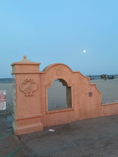 An outdoor scene depicts a decorative archway structure on a beach in Hollywood, United States of America. The archway, made of coral-colored material, features an arched opening looking out onto a sandy beach. A stylized "H" crest is visible on the left pillar of the archway, and a patterned tile is embedded on the right side. The sky above is a clear, pale blue with a full moon visible in the upper right quadrant of the image. The beach is populated with several people in beach attire, some sitting in chairs and others walking. The ocean is visible in the background. In the foreground, a concrete walkway with green painted lines is present. A sign with text in English and Spanish is attached to the left pillar of the archway, and some litter is scattered on the ground. The lighting suggests it is either dusk or dawn.