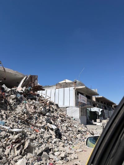 This image captures a scene in Jabalia, Palestinian Territory, depicting extensive urban damage under a clear blue sky. A large pile of concrete rubble, broken masonry, and twisted rebar dominates the left and central foreground, extending towards the right. Interspersed within this debris are various items, including dark and light-colored plastic bags and fabric scraps. To the right of the rubble, a multi-story building stands, showing signs of significant structural damage. Its lower facade is partially covered with white, vertically striped tarpaulin or sheeting. The upper sections of the building reveal exposed concrete and rebar, indicating partial collapse or heavy damage. A red and white sign, featuring Arabic text and a circular logo, is attached to a pole near an entrance at the building's ground level. This entrance area is further obscured by draped white canvas material. A thin, light-colored antenna or rod extends upwards from the building's roof. On the far right, a portion of a yellow vehicle's interior door frame and its exterior side mirror are visible; the mirror displays a crack and a distorted reflection. No individuals are present in the scene.