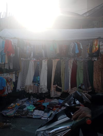 The image shows a street market stall in Kutawaringin, Indonesia (latitude: -6.974, longitude: 107.519). The sun is shining brightly, creating a hazy glare over the scene. A white awning shelters the stall, which is lined with rows of clothing items. A person's hand is visible, likely belonging to the vendor, resting on the front of a black scooter parked in front of the stall. There is an abundance of colorful clothing on display, and a variety of other merchandise is stacked and spread out on the ground in front of the stall. The image captures a typical scene of a bustling street market in a busy urban environment.