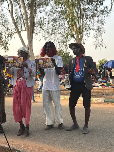 The image depicts three individuals in performative attire standing on an asphalt road in Lafia, Nigeria, alongside an open-air market under bright daylight.
From left to right:
1. A dark-skinned individual wears a white bonnet-like head covering, a light grey blazer over a checkered or plaid long-sleeved shirt with a red tie, voluminous pink trousers, and brown lace-up boots. White powder or paint is visible on the face. This person holds a glossy horizontal poster with both hands at waist level, displaying the title "ECHOES VICTORY" alongside multiple portrait photographs and the text "P.P.M" and "BALENCIAGA."
2. A second dark-skinned individual wears a black wig with red tips, dark eye makeup or paint, a plain white t-shirt, white loose-fitting trousers with perforated patterns, and grey slip-on shoes. This person also holds an identical "ECHOES VICTORY" poster horizontally with both hands at waist level, displaying the same content.
3. A third dark-skinned individual wears a straw hat, a dark blazer over a blue t-shirt featuring a white circular logo with "EF" or "LF" and "ECRA FAMILY," black shorts, and grey slip-on shoes. Red face paint or streaks are visible on the cheeks and forehead. A red bandana or scarf is tied around the neck. This person holds a silver-colored lantern in the left hand and gestures with the open right hand, palm forward, fingers spread.
The immediate foreground consists of asphalt road and a low green-painted concrete curb. Beyond the curb, reddish-brown soil is visible. The background features several tall, sparsely foliated trees against a clear, light sky. To the right and mid-ground, a bustling outdoor market is visible with various items, including textiles, spread on the ground. Several people are present, and portions of blue tarpaulin shelters are discernible.