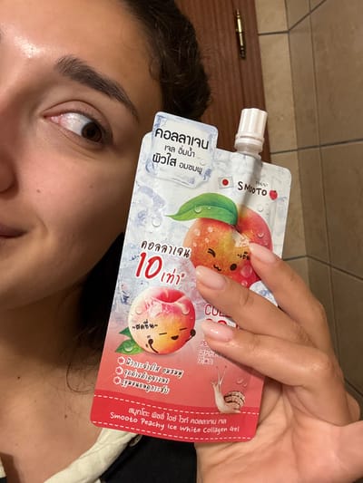 A female subject is partially visible from the mid-face upwards on the left side of the frame, smiling and looking directly into the camera. She has dark hair, dark eyebrows, and a brown eye. Her right hand, with manicured, natural-toned nails, holds a white, flexible pouch-packaging. The pouch features an illustration of peaches with cartoon faces, ice cubes, water droplets, and includes Thai script, the brand "SMooTO", a small Japanese flag icon, and the product name "Smooto Peachy Ice White Collagen Gel". The background consists of a light brown or beige tiled wall and a brown wooden door with a brass-colored hinge visible on the upper left. The specific location in Miljevci, Montenegro, cannot be determined from the visual evidence within the image.
