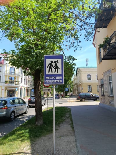 This media file depicts a street scene in Brest, Belarus, on a bright, sunny day. The focal point of the image is a blue traffic sign with a white border, featuring an illustration of a man and a woman embracing. Below this image, the sign reads "МЕСТО ДЛЯ ПОЦЕЛУЕВ" (Mesto dlya potseluyev), which translates to "Place for kisses." A smaller inscription below this, partially obscured, appears to be a date, "16.05.2019." This suggests the sign may mark a designated romantic spot.
The sign is positioned near a tree with green foliage and seed pods, casting dappled shadows on the grassy verge and a paved walkway. Several cars are visible on the street, including a silver SUV and a dark minivan. In the background, buildings with light-colored facades and balconies line the street. A pedestrian crossing is also visible, with a blue pedestrian crossing sign nearby. The sky is clear and blue, indicating pleasant weather. The overall atmosphere of the scene is calm and ordinary, with the prominent sign adding a touch of whimsy.