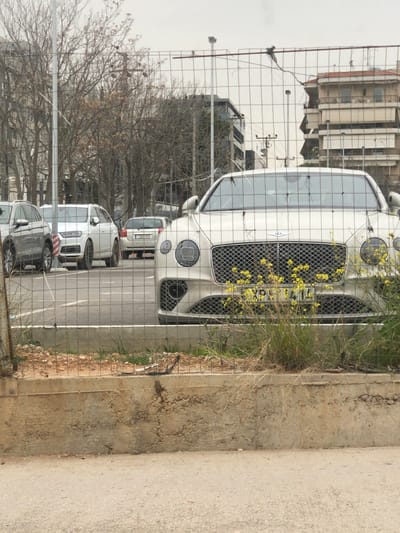 The image, rotated counter-clockwise for proper orientation, depicts a white Bentley Bentayga SUV prominently parked in an outdoor setting, viewed through a wire mesh fence. The luxury vehicle's large chrome grille, quad circular headlights, and front bumper are partially obscured by the fence and clusters of small yellow-flowering weeds growing alongside it.
Behind the Bentley, a line of other parked cars is visible, including a silver SUV or crossover and a darker vehicle further back. The background features several urban buildings, some appearing residential with balconies, and others more commercial or industrial in nature. Bare deciduous trees are scattered throughout the scene, suggesting either late autumn, winter, or early spring.
A concrete curb and an asphalt surface are present in the immediate foreground, beyond the wire fence. The lighting is diffuse, indicative of an overcast or hazy daytime sky, with no harsh shadows.
Visible text includes Greek license plates on the vehicles. The white Bentley's license plate displays "XP" followed by partially obscured numbers (e.g., "XP 110..."). A silver car further back also shows a Greek plate starting with "YNH". The overall scene captures a typical daytime view of parked cars in an urban or semi-urban environment, consistent with the provided location of Marousi, Greece.
