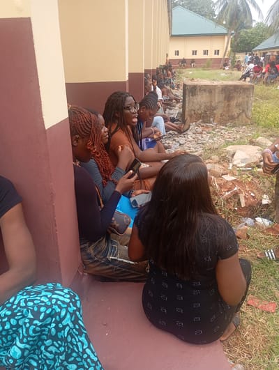 A medium shot taken in Ukwu-Abwa, Nigeria shows a group of people sitting on the ground along the exterior wall of a building. The foreground contains five individuals, who are all Black. The first woman is turned towards the camera, her face hidden, she has dark hair. Next, a woman with braided orange hair looks downward, holding a smartphone. Another woman, facing the same direction with dark braided hair and eyeglasses, has her mouth open as if speaking, and also holds a smartphone. Another woman sits near her, looking down at a phone as well. The fifth woman, closest to the camera, has long dark hair and is facing away from the camera. They are all sitting on the ground, against a wall painted brown and tan. In the background, across a small area of grassy and rocky ground, more people are sitting and standing. A building with a green roof and windows is visible in the background as well as a palm tree.