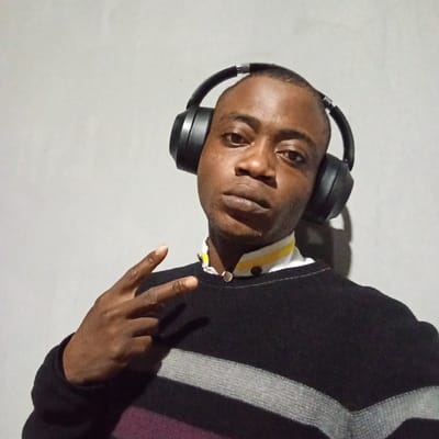 A young Black man with short hair, wearing black headphones and a black sweater with grey stripes, poses for a selfie against a plain grey wall. He is making a peace sign with his left hand. The image appears to be taken indoors, likely in a residential setting, given the plain backdrop and lighting. There is no indication of the specific location within Ibadan, Nigeria.