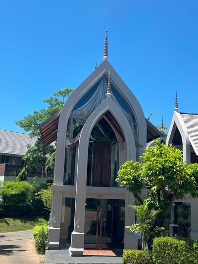 This is a vertical, eye-level shot of the exterior of a building with a unique architectural design, set against a clear blue sky. The building features a prominent, pointed roof structure with flowing, curved white beams that frame large glass windows, giving it a modern yet elegant appearance. The roof itself is tiled in a dark gray material.
Several pointed spires, resembling elongated pinecones with ribbed detailing, extend upwards from the white architectural elements. Lush green trees with dense foliage are visible around the building, some growing close to the structure, casting dappled shadows on the ground. One particularly large tree with bright green leaves is in the foreground, partially obscuring the view of the building's upper section.
To the right, a section of the building with wooden-slatted walls and a dark tiled roof is visible. In the lower part of the image, a pathway made of wooden planks leads towards the building, bordered by manicured green bushes and more trees. The sunlight suggests it is daytime, likely mid-morning or early afternoon, and the clear sky indicates fair weather.
No people are visible in the frame. The overall impression is one of serene, contemporary architecture integrated with nature in a tropical setting. The location context provided, Choeng Mon, Thailand, aligns with the tropical vegetation and architectural style, which often incorporates elements inspired by traditional Thai design but with a modern execution. No text is legible in the image.