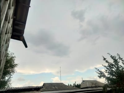 The image depicts a cloudy sky over a residential area. In the foreground, on the left, a portion of a weathered wooden structure with a dark-colored overhang is visible. To the right of this structure, several rooftops with corrugated metal and tile are present. A tall, thin pole with what appears to be a small antenna or object at its top stands on one of the rooftops. Green trees with full foliage are visible on the right side of the image, and a tree with sparser branches is seen in the center background. The sky is overcast with varying shades of gray and white clouds, with hints of lighter blue and pale yellow near the horizon, suggesting the presence of a sun obscured by clouds. The overall impression is a view from a window or elevated position looking out over a modest urban or village setting. The location in the city of Baş-Bulak, Kyrgyzstan, indicates a likely Central Asian environment.