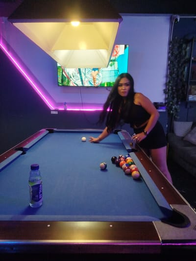 In Patong, Thailand, a woman is playing pool in a dimly lit establishment. She is wearing a black crop top and black shorts and has long, dark hair. The pool table is covered in a blue felt and has a rack of billiard balls set up on it. A white cue ball is positioned on the felt, and the woman is reaching out to strike it. A bottle of B'lue drink is placed on the table near the woman. The room is decorated with purple neon lights, and a large television screen is mounted on the wall. The atmosphere appears to be relaxed and casual, with the woman focused on her game.