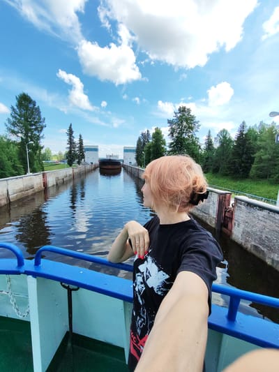 A medium shot captures a woman with light orange hair pulled back in a bun, wearing a black t-shirt with a graphic print, standing on the deck of a blue-railed boat. She is positioned on the right side of the frame, her face turned to the left, looking towards the water. Her left arm is extended, holding a phone to take a selfie. The boat's blue railing and white deck are visible in the foreground.
The background depicts a canal with dark, reflective water. On the left bank, a concrete wall lines the canal, with trees and a lamppost visible behind it. On the right bank, a similar concrete wall is present, with a metal lock mechanism visible. Further in the distance, a large cargo ship is slowly moving through the canal, flanked by two large, white, rectangular structures, likely part of a lock system. The sky above is a vibrant blue, dotted with fluffy white clouds, and bordered by a line of green trees on the far bank. The overall scene suggests a journey through a canal lock on a bright, cloudy day. The location is Belousovo, Russia, specifically within its canal system.