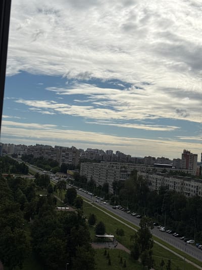 This is a rotated image of an urban landscape in Shushary, Russia. The scene is dominated by a tall, multi-story apartment buildings lining a street. The buildings are densely packed and appear to be of a uniform, somewhat older architectural style. A street runs alongside the buildings, with a row of parked cars visible along its edge. There is a sidewalk or pedestrian path next to the street, and beyond that, a grassy area with trees. The trees are lush and green, suggesting it is daytime during a warm season.

The sky occupies a significant portion of the image and is filled with dramatic, wispy clouds. These clouds are a mix of white and gray, with some areas appearing brighter where sunlight is filtering through. The overall lighting suggests it is either early morning or late afternoon, as the light has a slightly diffused quality. The weather appears to be partly cloudy, with patches of blue sky visible between the clouds.

The image captures a typical residential area in a Russian city, characterized by high-rise buildings and tree-lined streets. There are no people clearly visible in the image, and the focus is on the built environment and the sky. The perspective suggests the photo was taken from a high vantage point, possibly from a window of one of the apartment buildings. No text is discernible in the image.