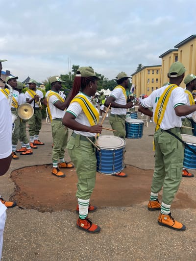 This media file depicts a group of young people participating in a parade in Alagba, Nigeria. The subjects are primarily individuals dressed in matching uniforms consisting of white shirts with green and yellow sashes, green trousers, green caps, and distinctive orange boots with green and white striped socks. Some individuals are holding drums of various sizes, with prominent blue snare drums visible. Others are playing brass instruments, including what appears to be a trumpet and a cymbal.
The scene is set on an asphalt road with a dirt embankment to the left. In the background, a light yellow building with multiple windows can be seen. The sky is overcast with clouds, suggesting it is daytime, but the lighting is somewhat diffused. The overall mood appears to be one of organized activity, possibly a musical performance or a ceremonial procession. There are hints of greenery from trees and possibly tents in the far distance, suggesting an outdoor event. One drum has the brand name "YAMAHA" partially visible. The sashes worn by some individuals have visible text, but it is largely obscured.