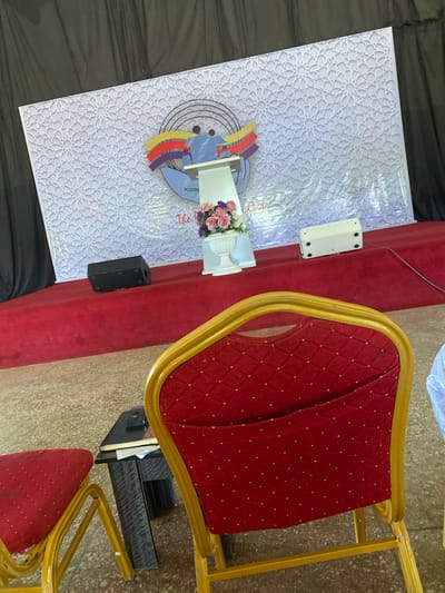 This image depicts an indoor scene, likely a stage or presentation area, set up with chairs and a backdrop. The backdrop features a white fabric with a light purple, repeating geometric pattern. Centered on the backdrop is a stylized graphic of two figures holding flags, with a lectern in front of them. A vase of pink and purple flowers sits to the right of the lectern.
In the foreground, there are several red chairs with gold-colored frames. Some of these chairs are partially visible, suggesting an audience seating arrangement. A small black table with books and a phone is situated between two chairs.
On a raised red carpeted platform behind the chairs, there are two audio speakers: one black rectangular speaker on the left and a white rectangular device on the right. The overall environment appears to be a venue prepared for an event. The mention of Barakin Lerawi, Nigeria, is context not visually evident in the image itself.