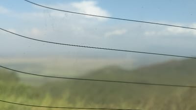 The image shows a blurred landscape viewed through a window with several horizontal defroster lines. The sky is light blue with some clouds, while the land features indistinct, grassy or bushy terrain. The view is partially obscured by smudges and minor imperfections on the glass surface, adding to the overall soft, diffused appearance.