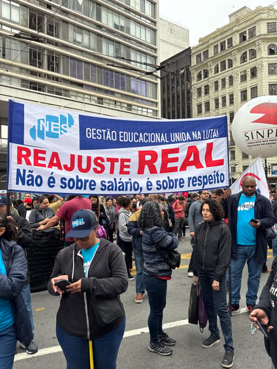 A street protest is taking place in what appears to be São Paulo, Brazil, where a group of people are marching, holding a banner with the inscription "GESTÃO EDUCACIONAL UNIDA NA LUTA!" (Educational Management United in the Struggle!), "REAJUSTE REAL" (Real Adjustment), and "Não é sobre salário, é sobre respeito" (It's not about salary, it's about respect). A SINPE balloon is visible. People are wearing casual clothes and holding phones, while some are looking at the banner and walking. Buildings form the background.