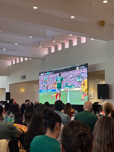 The image depicts an indoor gathering of people watching a soccer match on a large screen in a venue that appears to be the "Estadio de Béisbol Hermanos Serdán" in San Andrés Cholula, Mexico. The screen displays a match with players in green and yellow uniforms. The crowd is composed of individuals of various ages, many wearing green shirts, suggesting support for the Mexican team. The environment includes a white ceiling with recessed lighting, and speakers are visible on either side of the screen. The overall atmosphere suggests a communal viewing experience of a sporting event.