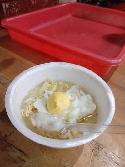 The image captures a close-up view of a bowl of instant noodles, set in what is likely Pangalengan, Indonesia. The central subject is a white, round styrofoam bowl containing cooked instant noodles submerged in a light-colored broth. Floating in the broth are two distinct pieces of egg: a fully formed, soft-boiled or poached yellow yolk, and a separate portion of cooked egg white. A clear plastic spoon is partially submerged in the broth, positioned towards the front of the bowl.
The bowl rests on a wooden surface, which appears to be a table or counter, characterized by its light brown color and visible grain and imperfections. In the background, out of focus, is a large, rectangular, bright red plastic tray or container. The lighting suggests an indoor setting, possibly with artificial light, given the shadows visible on the wooden surface. There are no people visible in the frame, and no discernible text. The scene depicts a simple, ready-to-eat meal.