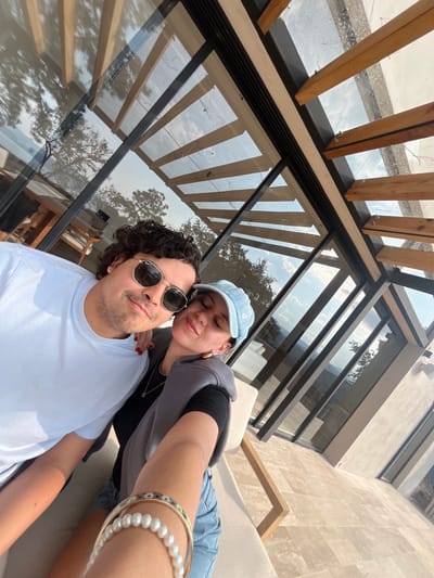 A couple poses for a selfie outdoors. The man wears sunglasses and a light blue shirt, while the woman dons a denim cap and gray hoodie. They are positioned against a background featuring a modern building with large glass windows and a wooden pergola above. Pearl and gold bracelets adorn the woman's wrist.