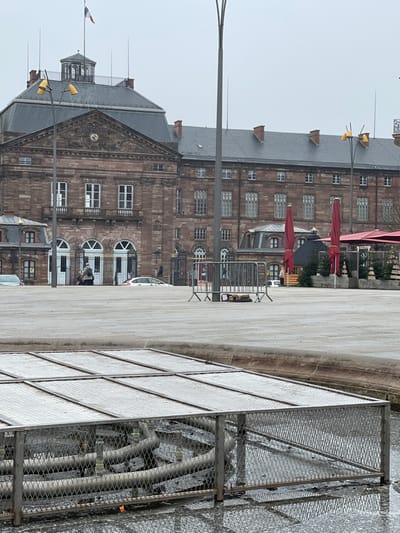 The image depicts an overcast daytime scene in Saverne, France, focusing on the Château des Rohan and its adjacent public square. In the immediate foreground, a rectangular, dormant fountain structure is covered by multiple panels of silver-colored metal grating, with standing water visible within and around its base.

The midground features an expansive, gray paved square. Two tall, dark gray streetlights with multiple yellow lamp heads rise vertically from the pavement. Several silver-colored temporary crowd barriers are positioned across the square. Two individuals are discernible near the main arched entrance of the Château des Rohan, with a white sedan partially visible behind the barriers. Another individual is faintly visible further to the right, near the market area.

The Château des Rohan, a prominent multi-story building constructed of reddish-brown stone, forms the background. Its facade features a central pediment containing a clock, numerous rectangular windows with white frames, and grand arched main entrances with white double doors behind ornate black gates. A dark gray slate roof with several chimneys covers the structure, and a French flag flies from a cupola on the roof. To the right of the Château, several red closed patio umbrellas, a wooden market stall resembling a chalet, and small decorated Christmas trees are arranged, indicating a seasonal outdoor display or market setup.