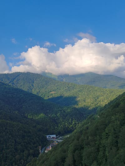 This is a landscape photograph taken in Estosadok, Russia. The image captures a vast mountainous region covered in dense green forest. Rolling hills and prominent mountains fill the mid-ground and background, with the slopes of the mountains in the foreground also heavily forested. The sky above is a clear blue, interrupted by a large, bright white cloud formation.

In the valley between the mountains, a small settlement is visible. This settlement appears to be a village or town, with several buildings, including what looks like a church with a steeple, and other colorful structures. A tall tower, possibly a communication tower or part of a ski lift, stands near the village. A road also winds through the settlement and into the forest.

The lighting suggests it is daytime, with sunlight casting shadows across the mountains, creating a dynamic interplay of light and shade. The lush greenery indicates a temperate climate during a warmer season. The overall scene evokes a sense of tranquility and natural beauty. There is no visible text in the image.