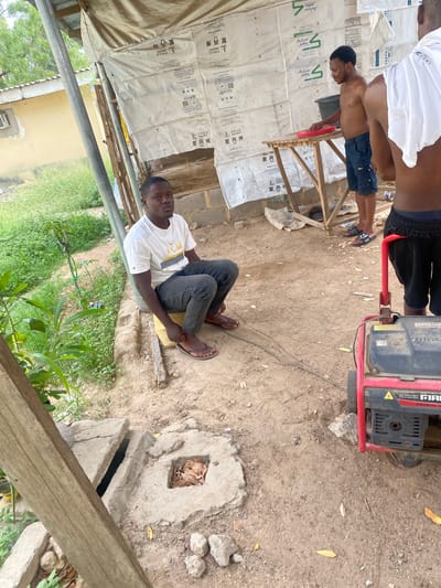 The image depicts an outdoor scene in Giginyu, Nigeria, likely within a construction or workshop area. In the foreground, a man is seated on a rough yellow mat, wearing a white t-shirt with "ACM" printed on it and dark grey jeans. He is looking towards the viewer. To his right, a red portable generator is situated on the sandy ground. Behind the seated man and to the right, another man, shirtless and wearing dark blue shorts, stands near a wooden table holding a red object. A third man, partially visible from the back and wearing a white t-shirt, stands to the far right of the frame. The background features a structure with a tarp-like covering and exposed brickwork, with debris scattered on the ground. Lush green vegetation is visible on the left side of the image. The environment appears unkempt, with evidence of construction or ongoing work.