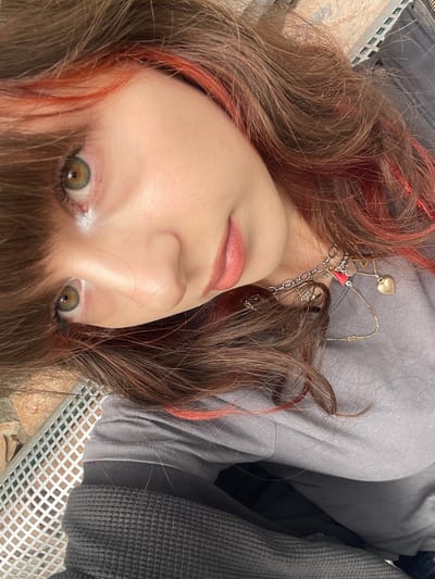 This image, taken in Raanana, Israel, presents a close-up, diagonally oriented portrait of a female individual. Her head and upper torso are visible. She has medium brown hair with distinct reddish-orange highlights, styled with bangs across the forehead. Her large eyes, appearing hazel or green-brown, gaze directly forward. Her skin is fair, with a slight sheen on the upper cheekbone, and her lips are a natural pink tone, slightly parted. She is wearing a gray short-sleeved t-shirt, beneath which a dark gray waffle-knit garment is visible. Layered necklaces are present around her neck, consisting of a silver-toned chain with multiple charms, including a small red rectangular pendant and a blue 'evil eye' bead, along with gold-toned chains featuring a heart-shaped pendant and a "V" shaped pendant. The background, visible to the lower left, consists of a light-colored, reddish-brown stone surface and a metallic, light-colored grid structure, indicative of an outdoor setting.