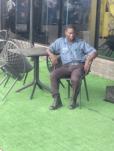 The image displays an outdoor seating area in Ikeja, Nigeria. A male individual is seated, facing forward, on a dark grey metal chair featuring an open geometric pattern on its back and seat. He is dressed in a light blue short-sleeved collared shirt with a "SECURE NIGERIA" patch on the left chest and a blue belt. He wears dark brown trousers, a black combat boot on his left foot, and a black dress shoe on his right foot. A thin blue stripe runs vertically down the outside of his right trouser leg. His arms rest on the chair's sides, and he maintains direct eye contact with the camera. To the left of the seated individual is a dark grey rectangular table with a central pedestal base branching into four legs. Another similar dark grey metal chair is partially visible to the left of the table. The ground surface consists of green artificial turf. In the background, a building with large glass windows reflects multiple indistinct figures and urban elements. A yellow sign, partially showing "CRAPES CH", is discernible through or behind the glass on the right side.