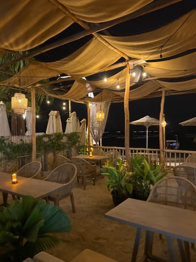 The image depicts an outdoor restaurant or seating area at night, likely on a beach or near a body of water. The location is specified as Unknown, Indonesia. The scene is illuminated by string lights and lanterns, casting a warm glow on the sandy ground, woven chairs, and wooden tables. Overhead, a canopy of tan fabric is supported by bamboo poles, creating a rustic and bohemian ambiance. In the background, the dark expanse of water is visible, with a few distant lights suggesting a shoreline or boats. Several white beach umbrellas are stacked and covered, indicating the restaurant may be closed or preparing for the day. Lush green plants are strategically placed in pots, adding a touch of nature to the setting. The overall atmosphere is serene and inviting, suggesting a place for relaxation and dining.