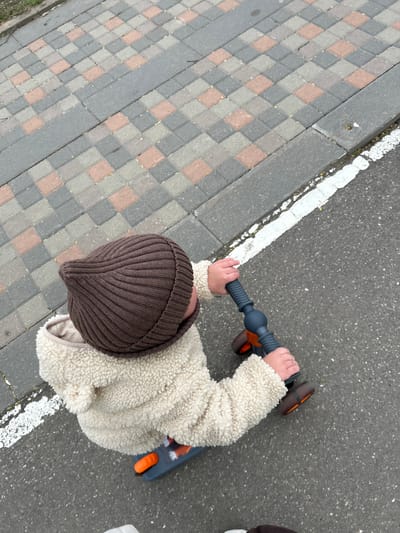 A top-down shot shows a young child in a brown knit hat and a cream-colored fleece jacket standing on a paved surface next to a section of multicolored paving stones. The child is holding the handlebars of a blue and orange scooter. The paved surface appears to be a street or sidewalk, with a white line marking its edge. The paving stones are arranged in a geometric pattern with gray, brown, and light gray hues. The scene suggests an urban environment, potentially in Klin, Russia.