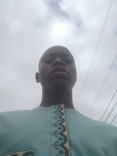 A low-angle, close-up shot of a dark-skinned young man wearing a light blue shirt with decorative trim. He is centered in the frame with his head tilted slightly back. The background is overcast, featuring gray clouds. Several utility wires are visible against the cloudy sky in the upper right corner of the frame. The scene is possibly located in Epe, Nigeria.