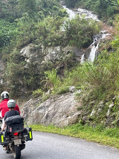 The image captures a scenic moment in Tân Quang Commune, Vietnam, featuring two individuals on a motorcycle stopped beside a winding asphalt road, observing a lush mountain landscape. The motorcycle, an adventure-style model, is equipped with black saddlebags or panniers on either side, which have bright yellow reflective strips. A red taillight is visible at the rear.

Two people are seated on the motorcycle. The rider wears a bright red jacket and a light grey helmet, while the passenger behind them is dressed in a dark grey or black jacket and a dark helmet, with a backpack visible between them. Both appear to be looking towards the left side of the frame, indicating they are taking in the view.

To the left of the road, a steep, rocky mountainside dominates the scene. It is densely covered with various shades of green vegetation, including tall grasses, bushes, and ferns, thriving on the rock faces. A prominent white waterfall cascades down the mountain in several sections, creating a dynamic visual element against the dark, wet rocks and vibrant greenery. The lighting suggests it is daytime, with soft, diffused light, possibly indicating an overcast sky, as there are no harsh shadows. The scene conveys a sense of a journey through a rich natural environment. No legible text is visible in the image.