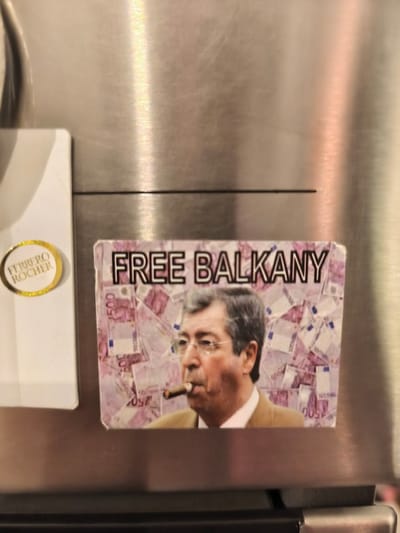 The image depicts a sticker on a stainless steel refrigerator surface. The sticker features a man with a graying beard and hair smoking a cigar and wearing a white shirt and gray suit jacket. The sticker is filled with Euro bank notes. Above the man, the text "FREE BALKANY" is displayed in black outlined white font. The sticker is located on a stainless steel refrigerator in a residential building in the city of Saint-Josse-ten-Noode, Belgium at the coordinates (latitude: 50.849, longitude: 4.378). A white card is to the left of the sticker that displays a gold-bordered circular logo, with text "FERRERO ROCHER". The sticker is affixed to the refrigerator at a slight angle. The refrigerator is located in a home or apartment.