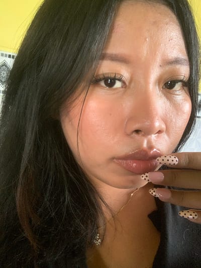 A close-up, waist-up shot shows a young woman with long, black hair. She is looking down and to her right, her mouth slightly open. Her lips are glossy and her eyes are dark. She has long, false eyelashes and her eyebrows are well-defined. Her skin appears to be slightly oily. She is wearing a gold chain necklace with a small, sparkling pendant. Her right hand is raised to her face, with her fingernails painted with a light pink base and black polka dots. The background is blurred, but appears to be indoors with a yellow wall on the left and a patterned surface on the right. The specific location in the city of Jajag, Indonesia cannot be determined from the visual information provided.