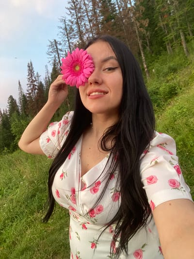 In a grassy field in the city of Olkhovka, Russia, a young woman with long, dark hair is captured in a selfie. She is wearing a white dress adorned with pink roses. A bright pink gerbera daisy is held over her right eye, obscuring it from view. Her left eye is visible, and she is smiling, revealing her teeth. Her left arm is bent, with her hand holding the flower to her face. The background consists of a dense forest with tall, slender trees and a partly cloudy sky. The lighting suggests it is either early morning or late evening.
