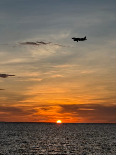 A silhouette of an airplane is captured against a twilight sky over the Gulf of Thailand, near Choeng Mon, Thailand. The sun is setting, casting an orange and yellow glow across the sky and the calm ocean surface. The airplane, a commercial jet with landing gear extended, is in the upper right quadrant of the image, appearing to be on its descent. The water is dark and textured with gentle ripples. The horizon line is clearly defined between the sea and the sky, which transitions from pale blue at the top to vibrant oranges and yellows closer to the horizon. Scattered clouds are present in the sky, some tinged with the colors of the sunset.
