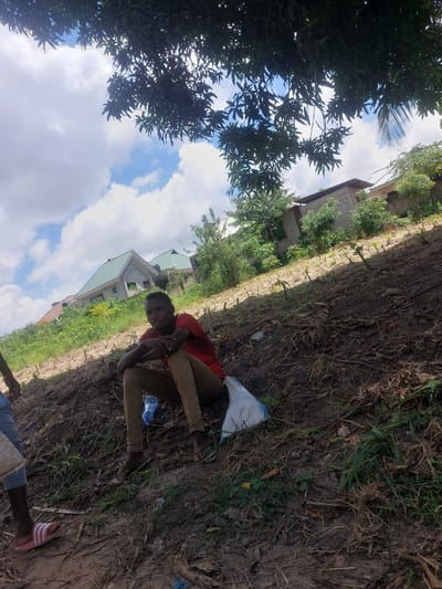A young person wearing a red shirt and tan pants sits on the ground. Another person's legs are partially visible near the top-left of the frame. A white and blue plastic bag and a plastic water bottle are near the seated person. The ground appears to be dirt and dry leaves. A row of small plants or trees stretches across the ground behind the seated person. In the background are buildings and trees under a partly cloudy sky. The scene could be in an agricultural or residential area of Dar es-Salaam, Tanzania.
