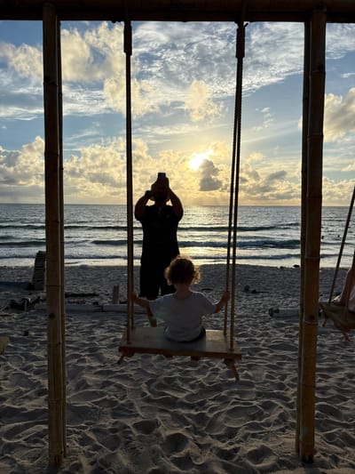 This media file shows a father and son on a beach in Choeng Thale, Thailand. The father is silhouetted against the setting sun, holding up his phone to take a picture. His son is sitting on a wooden swing, looking out at the ocean. The sand is rippled, suggesting the tide has recently receded. The sky is filled with dramatic clouds, illuminated by the low sun. The overall mood is peaceful and serene, with a hint of joy in the father's action. The image is framed by bamboo structures, likely part of a beach swing set or cabana. The time of day is clearly late afternoon or early evening, as indicated by the low angle of the sun and the golden light. The weather appears to be clear with scattered clouds.