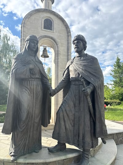 The image shows a bronze monument of Prince Peter Muromsky and his wife, Princess Fevronia of Murom, standing in front of an archway structure with a bell hanging inside. Both figures are clad in traditional medieval Russian clothing. The prince is wearing a cape and holds a sword. The statue stands in the city of Dmitrov, Russia, as determined by the surroundings in the background. The sky is visible behind the statues and includes blue sky and clouds. Green trees and grass are visible behind the monument. The statue is on a grey stone base.