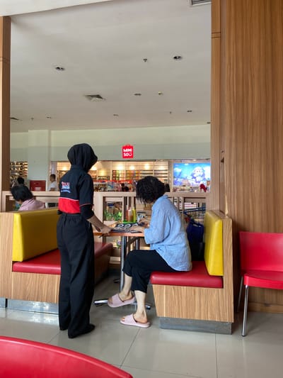 The image depicts the interior of a brightly lit, modern cafe in Kudus, Indonesia. A female server wearing a black headscarf, black pants, and a black and red uniform shirt stands to the left, holding a menu. She is interacting with a female customer who is seated at a table. The customer, dressed in a light blue shirt and black pants, is looking down at something on the table. The cafe features booth seating with red and yellow cushions, and wooden paneling. In the background, a store called "MINI SO" is visible, along with shelves stocked with merchandise. Another customer is seated at a booth in the background, to the left of the server. The floor is tiled and reflects the light.