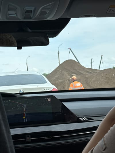 The image, taken from inside a vehicle, looks out towards a roadside scene under an overcast daytime sky. The perspective is from the passenger side, with interior elements framing the view. On the far left, a grey car sun visor with a "no airbag" warning symbol is visible. Below it, part of the car's dark grey interior paneling is seen. On the right, a large digital display shows a navigation map with a route highlighted in red and yellow, indicating a speed limit of "50" in a circle and a number "6" (possibly indicating time or distance). A person's arm or shoulder is also visible on the far right.

Outside the vehicle, a construction or earthworks site is visible, dominated by a large brown dirt mound on the right. A construction worker wearing an orange high-visibility vest and a light-colored hard hat or cap stands near the dirt mound. Several tall, slender poles or gantries extend into the sky behind the mound and in the distance. Ahead of the viewing car, partially obscured by the car's interior, is the rear of a white BMW X6 SUV, with its "X6" badge and circular BMW logo clearly visible. The overall scene suggests travel through an area undergoing construction or industrial activity, possibly in Kabanikha, Russia, as indicated by the context.